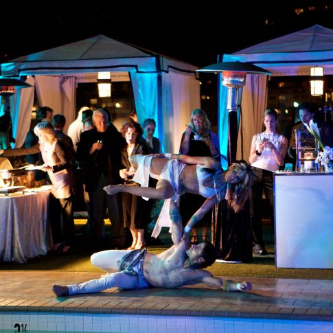 Professional acrobatic performers entertaining guests poolside during a luxury private event with elegant cabanas and evening lighting.