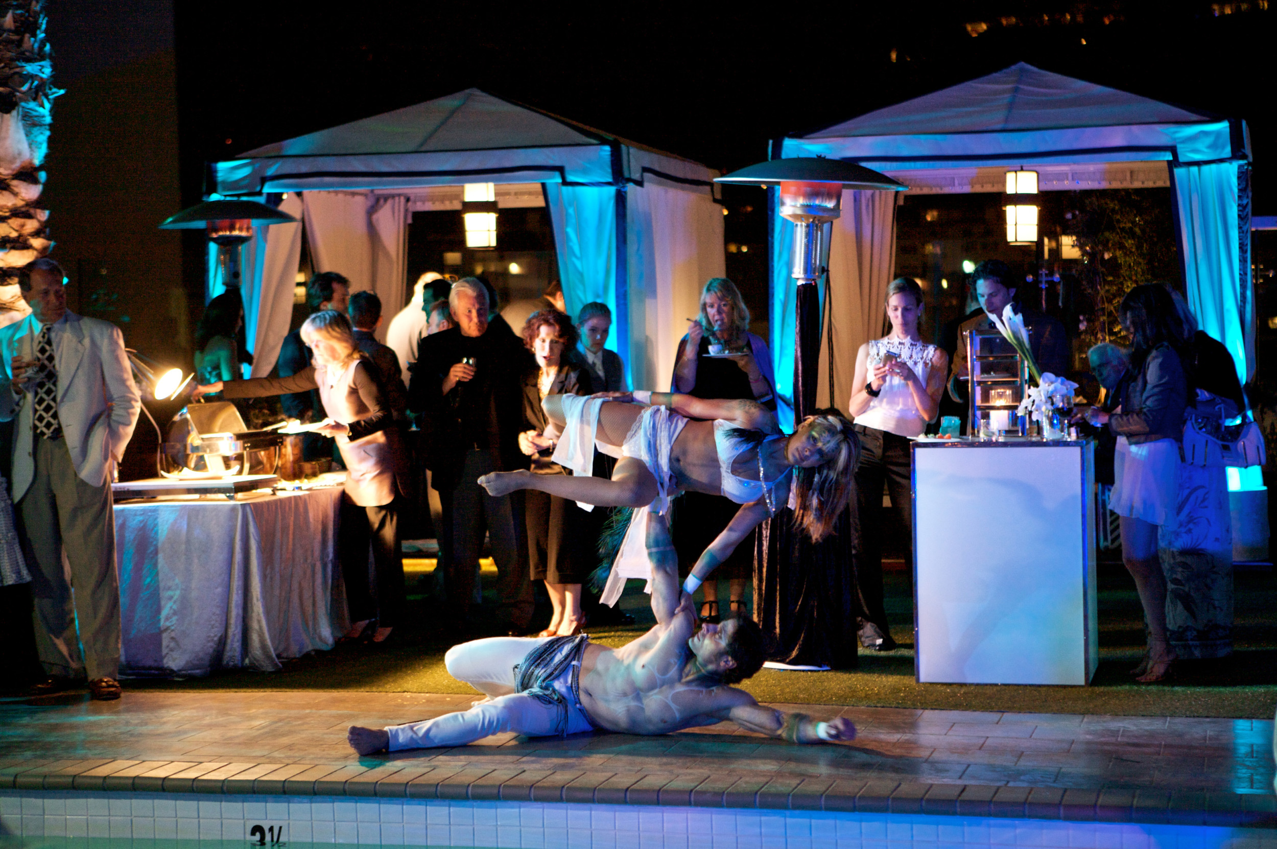 Professional acrobatic performers entertaining guests poolside during a luxury private event with elegant cabanas and evening lighting.