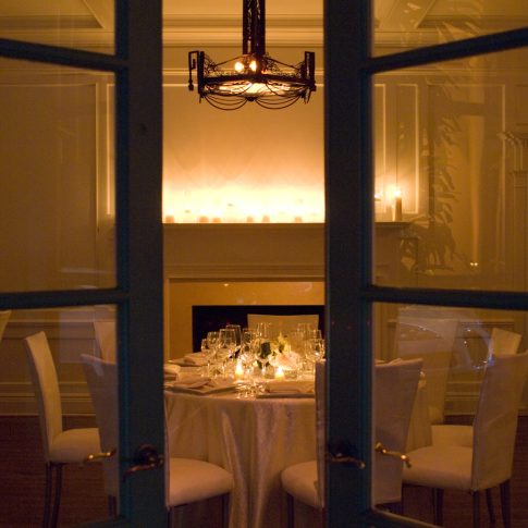 Elegant candlelit dinner table inside a luxury private estate, viewed through French doors with a fireplace, warm lighting, and intimate evening ambiance.