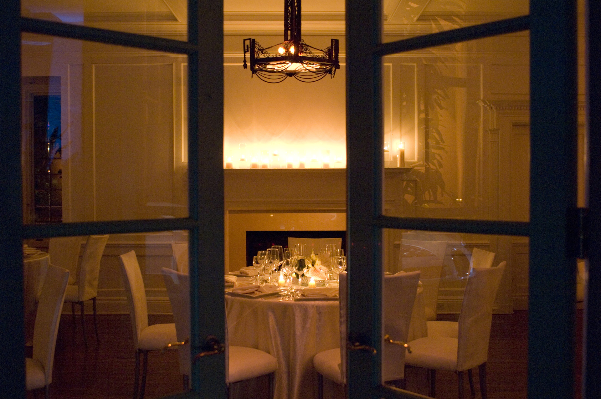 Elegant candlelit dinner table inside a luxury private estate, viewed through French doors with a fireplace, warm lighting, and intimate evening ambiance.