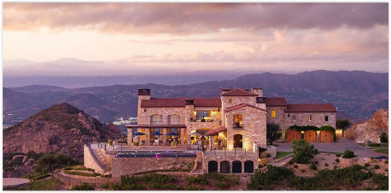 Luxury Malibu vineyard estate at sunset featuring Tuscan-inspired architecture, an infinity pool, rolling vineyards, and panoramic mountain views